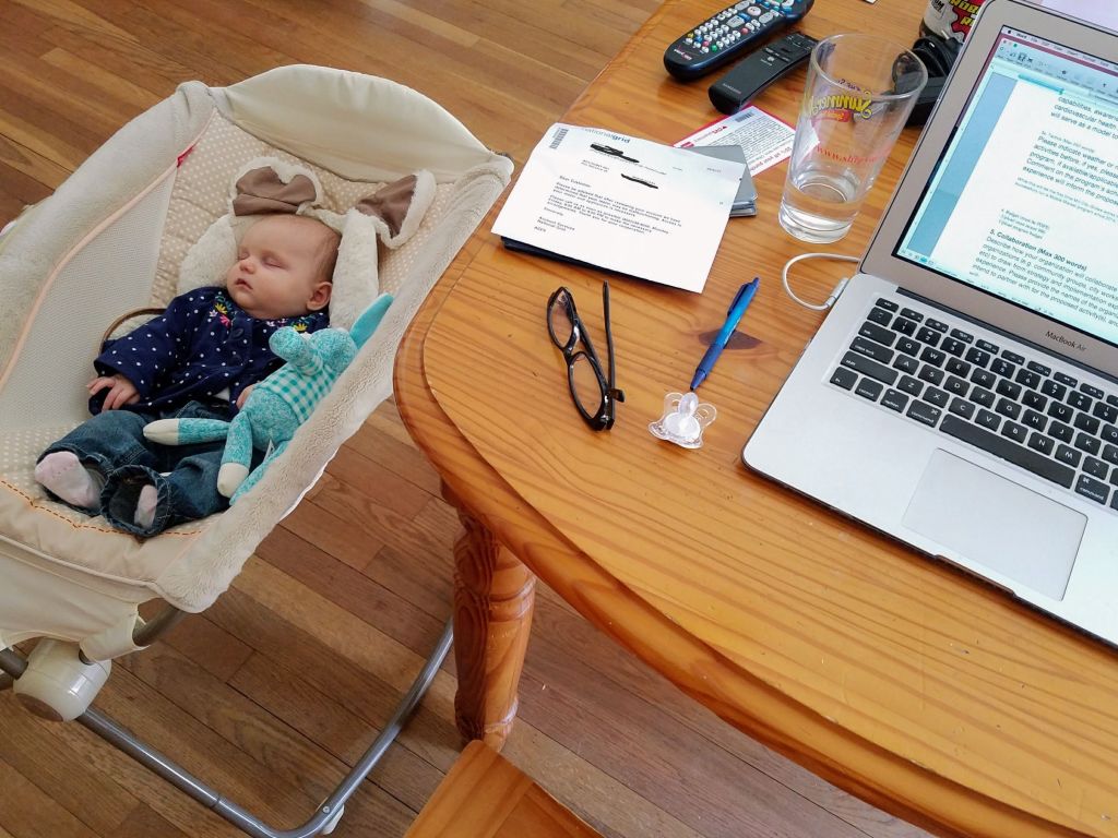 a baby sleeping next to a desk where glasses, papers and an open laptop sit. Presumably an adult is working while the baby is taking a nap.